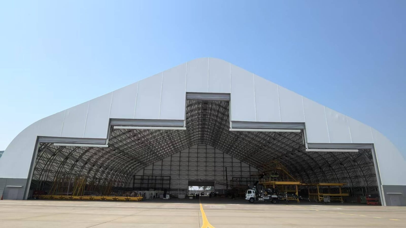 Large white hangar with a curved roof, open entrance revealing steel framework and equipment under a clear blue sky.