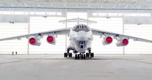 Front view of a large four-engine jet in a spacious hangar, red covers on its engines.