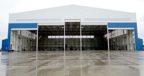 Large white and blue aircraft hangar with open doors and an empty interior, a wet concrete apron in front.
