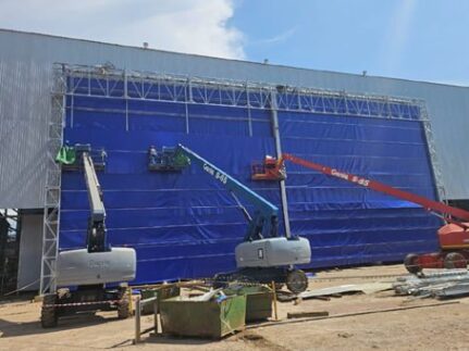 Construction site workers on elevated platforms install a large blue temporary facade on a warehouse wall behind scaffolding and equipment to the side, daytime sunlight.