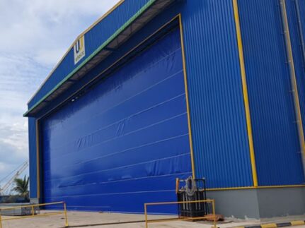 Blue industrial warehouse with a large blue roll-up door and yellow trim in an outdoor loading area.",