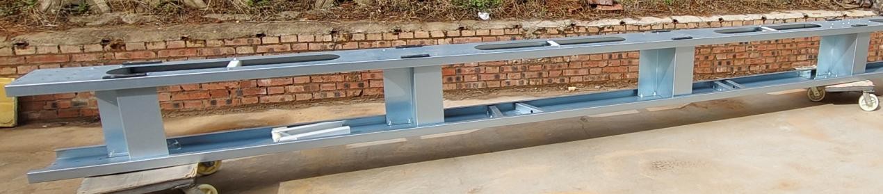 Long blue steel beam with rectangular support bays on casters, resting on a concrete floor against a brick wall (industrial equipment)