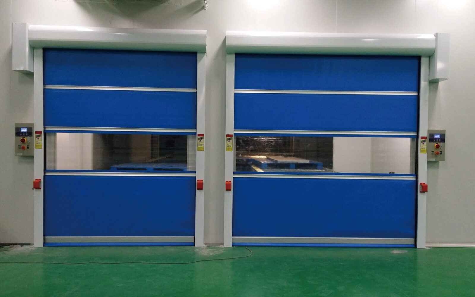 Pair of blue industrial doors in a warehouse, closed, with white framing and control panels on both sides over a green floor.
