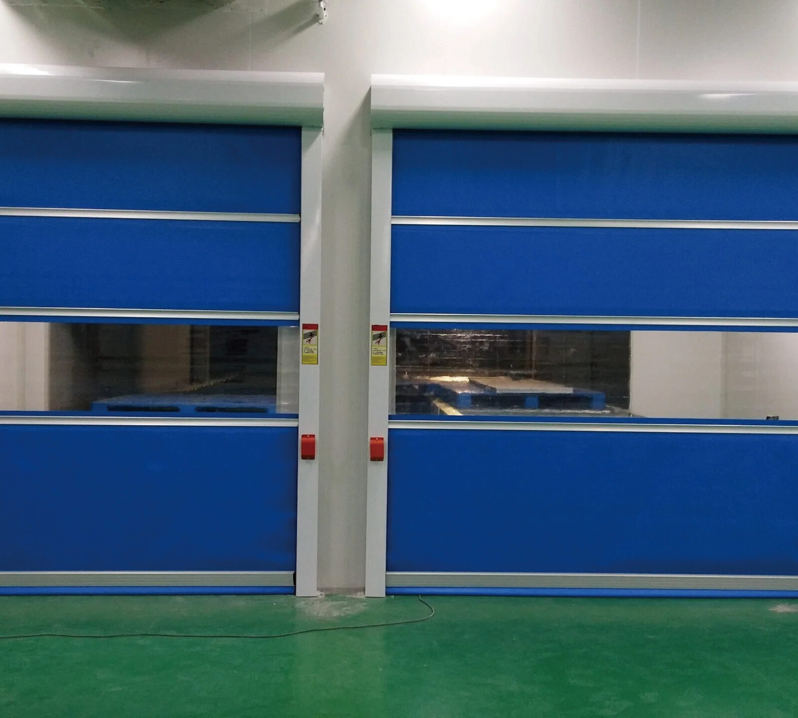 Pair of blue industrial doors in a warehouse, closed, with white framing and control panels on both sides over a green floor.
