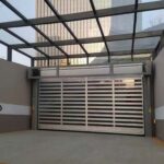 Industrial car entry with a wide metal rolling gate, gray walls, and a glass-framed overhead structure.
