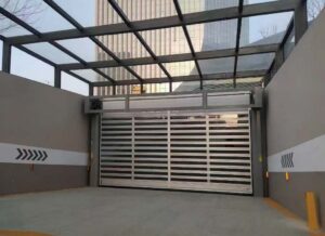 Industrial car entry with a wide metal rolling gate, gray walls, and a glass-framed overhead structure.